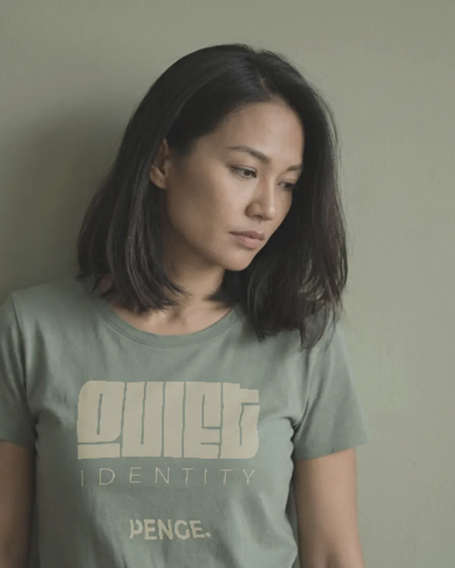 Model in the sage QUIET IDENTITY tee, looking down, against a green wall